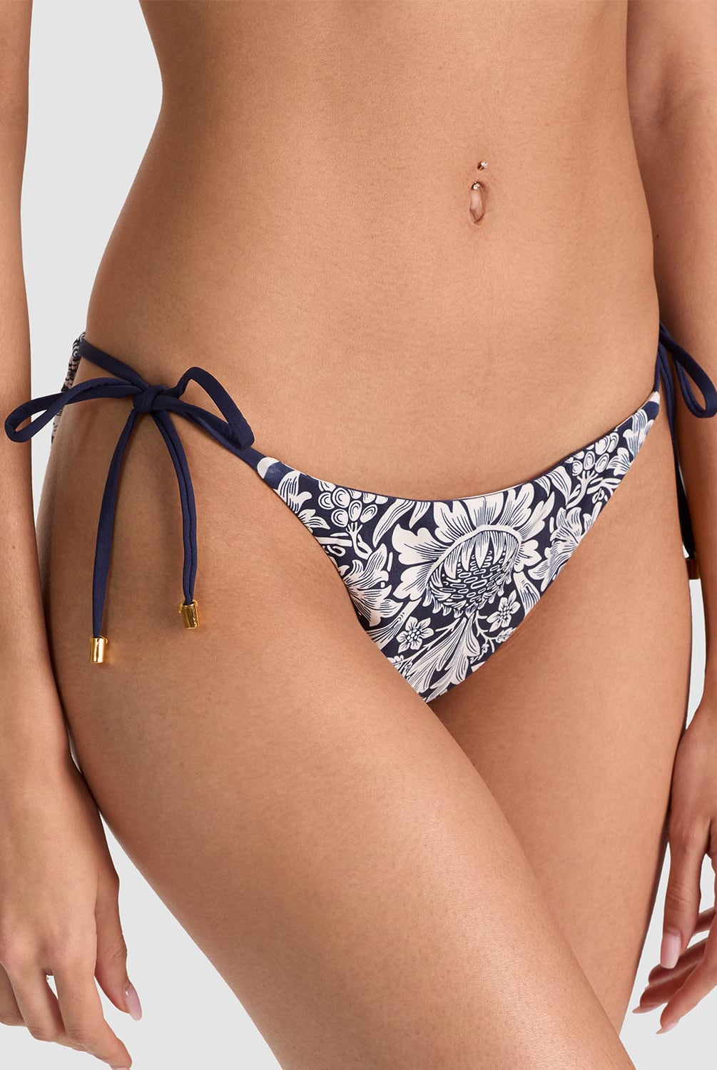 A close-up of a model wearing bikini bottoms with a navy blue and white floral pattern, featuring side-tie straps with gold-toned tips, against a light gray studio background.