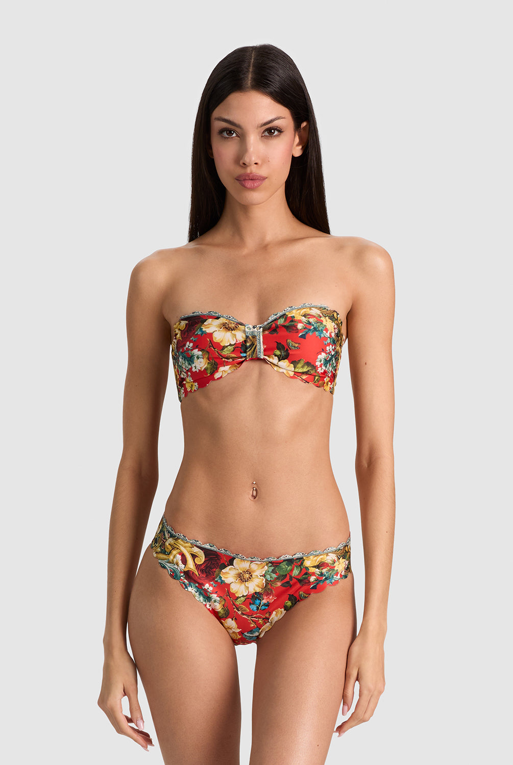 A model wearing a strapless bandeau bikini with a vibrant red, yellow, and green floral pattern, set against a solid light gray background.