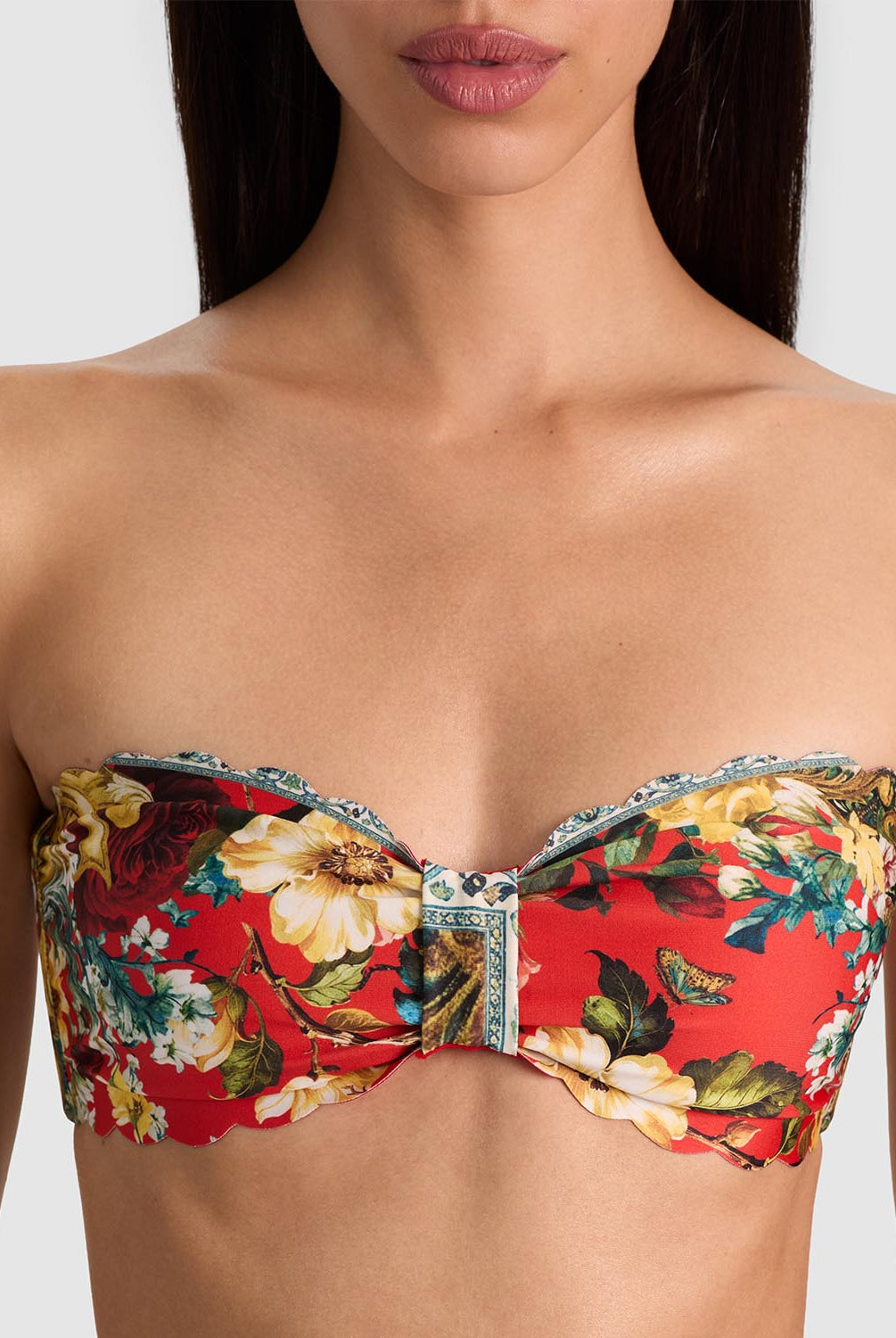 A close-up of a model wearing a strapless bandeau bikini top with a vibrant red background and a detailed floral print in yellow, green, and white, featuring a central decorative knot.