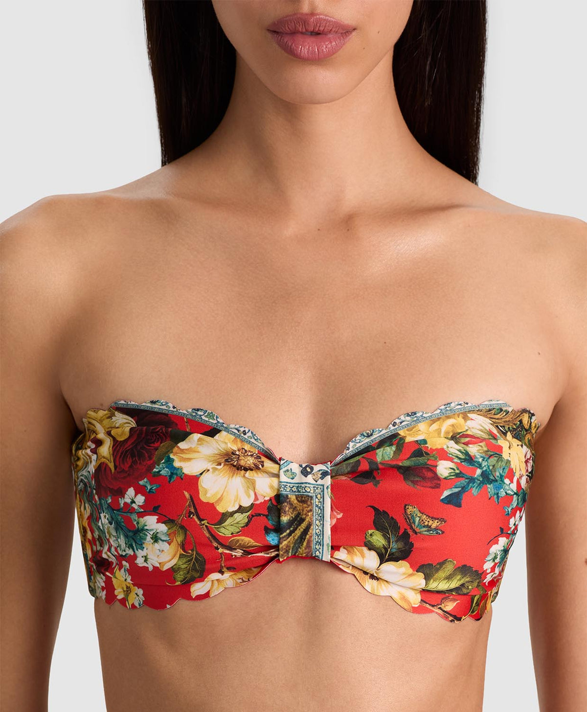 A close-up of a model wearing a strapless bandeau bikini top with a vibrant red background and a detailed floral print in yellow, green, and white, featuring a central decorative knot.