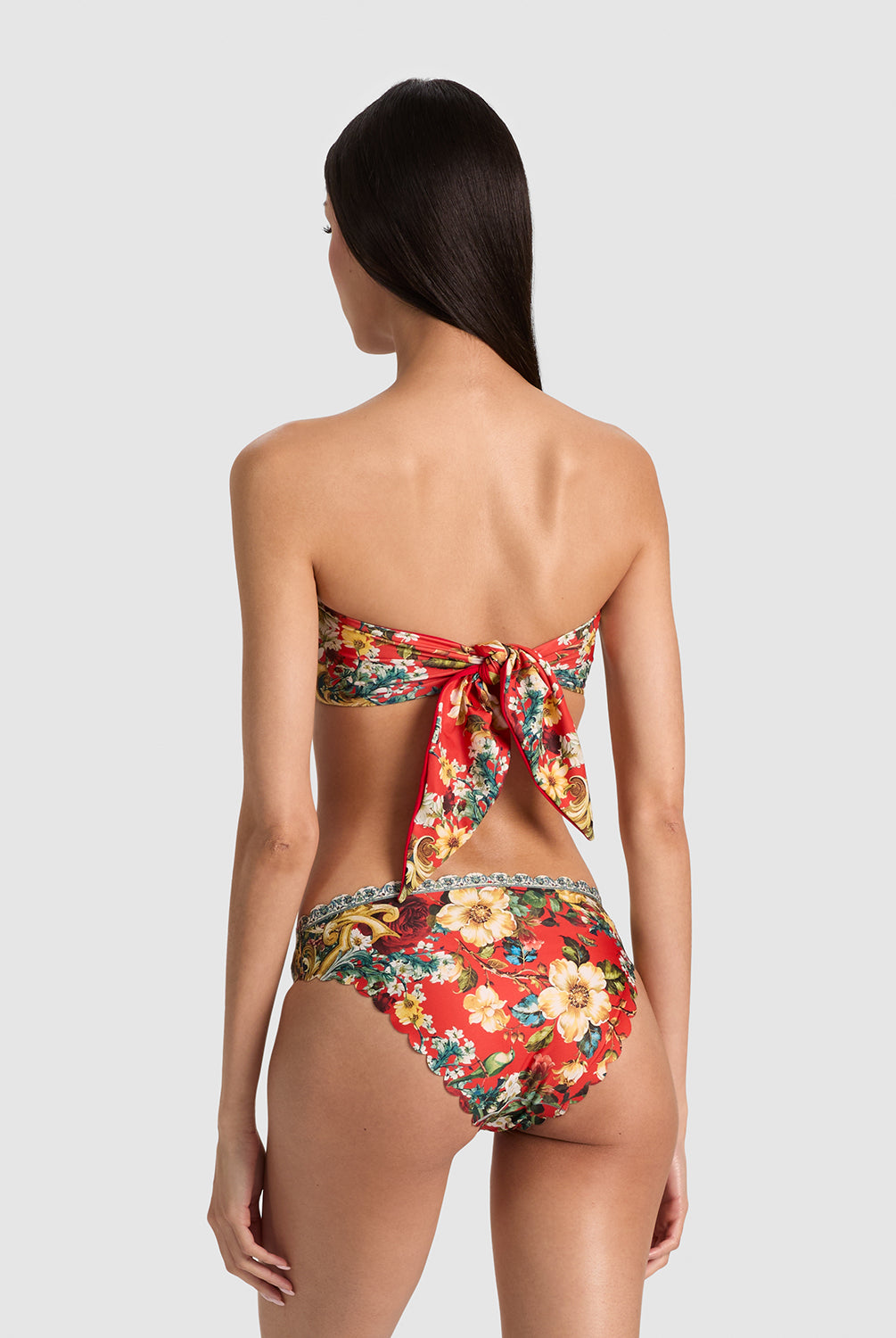 A woman shown from the back wearing a strapless floral bikini with a large bow detail at the back of the top and high-cut bottoms, against a light gray studio background.