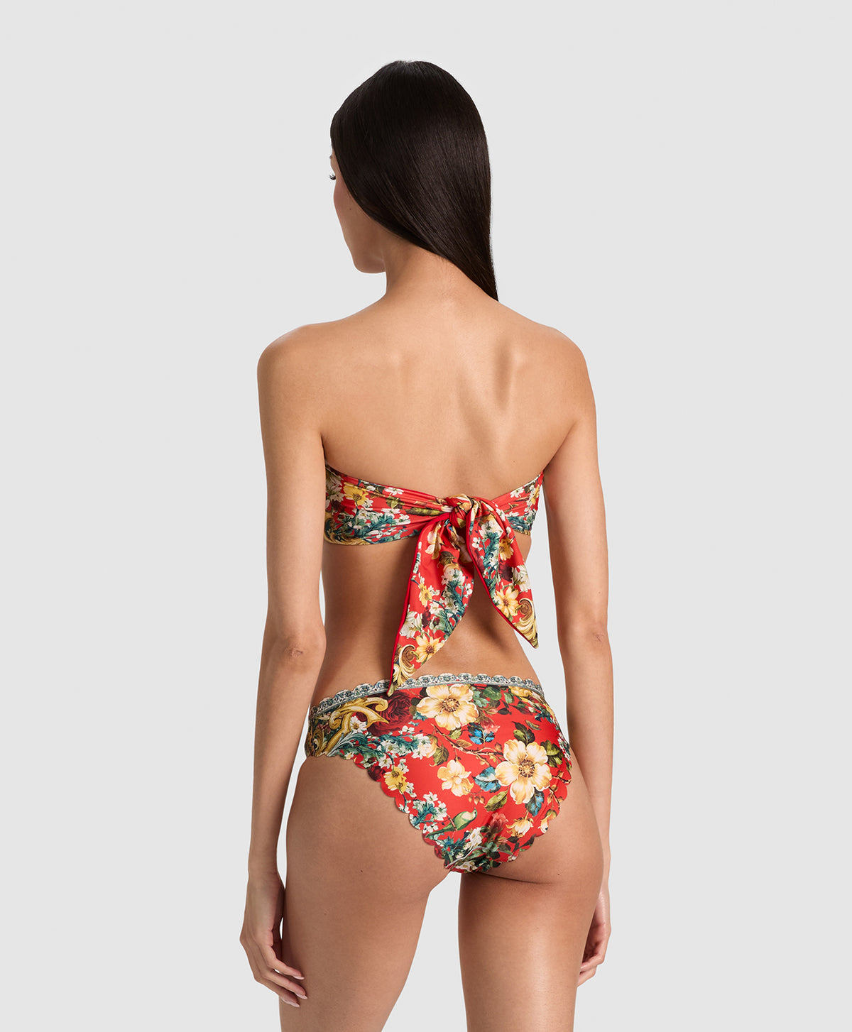 A woman shown from the back wearing a strapless floral bikini with a large bow detail at the back of the top and high-cut bottoms, against a light gray studio background.
