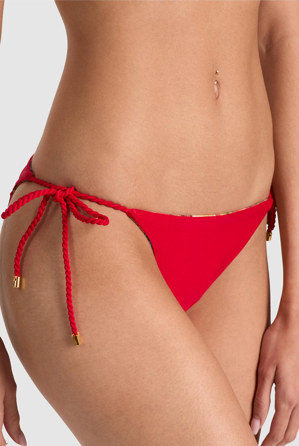 A close-up of a model wearing bright red bikini bottoms with side-tie rope details and small gold hardware accents, photographed against a light gray studio background.