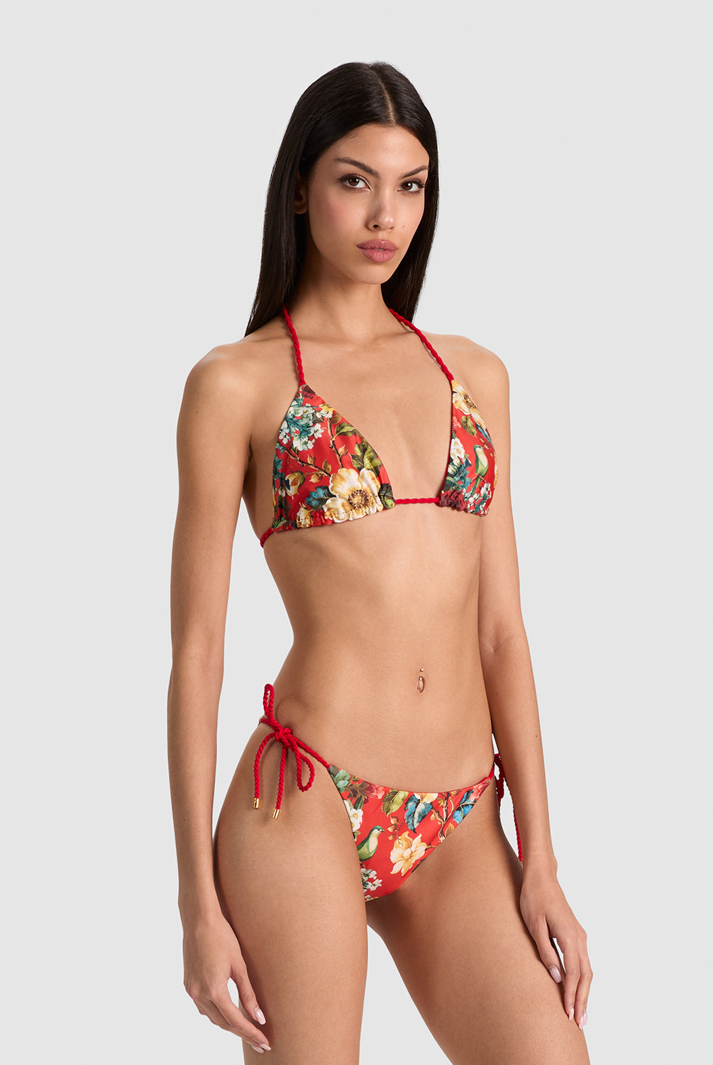 A woman modeling a two-piece bikini with a red string base and a colorful floral print, photographed against a solid light gray background.