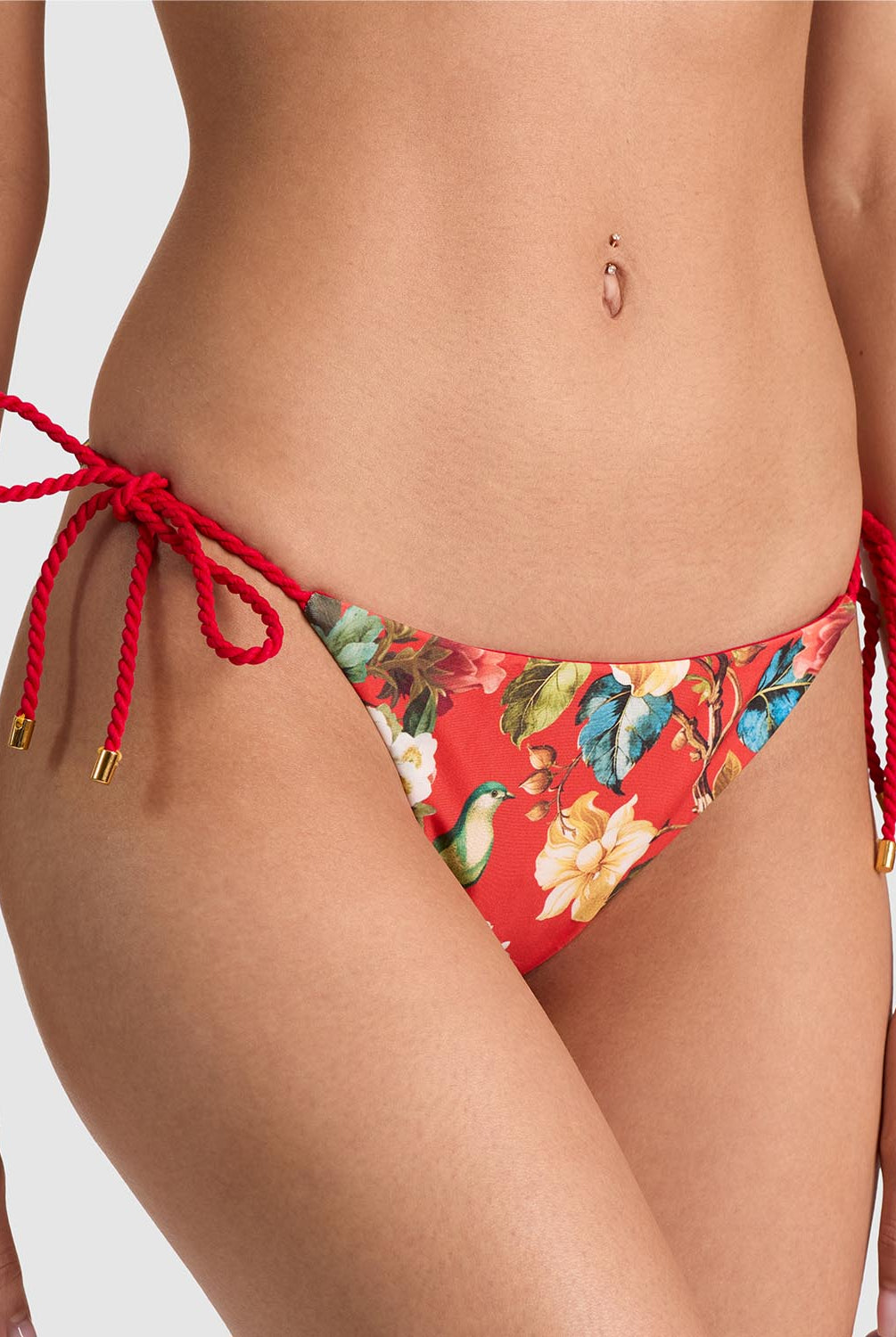 A close-up of a model wearing red bikini bottoms with a tropical floral print and red rope side ties with gold-toned tips, shown against a light gray studio background.