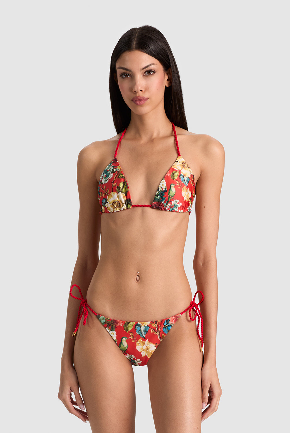 A woman modeling a two-piece string bikini with a red base and a multicolor floral print, set against a solid light gray background.