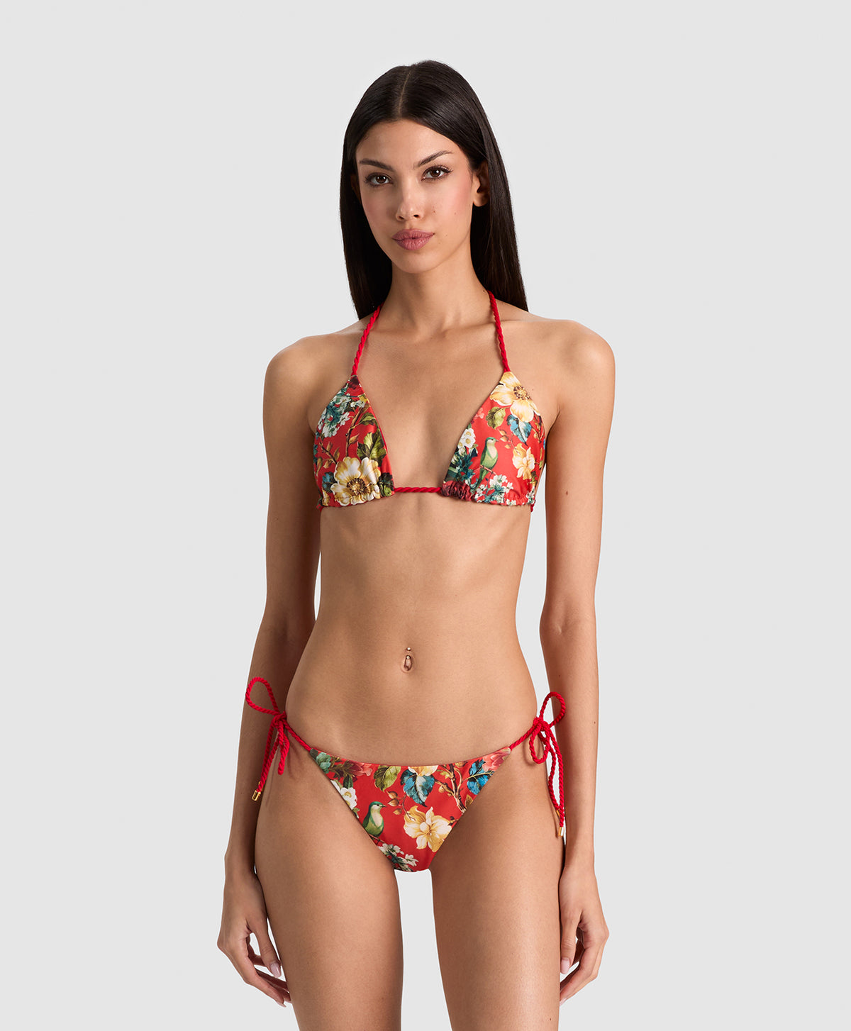 A woman modeling a two-piece string bikini with a red base and a multicolor floral print, set against a solid light gray background.