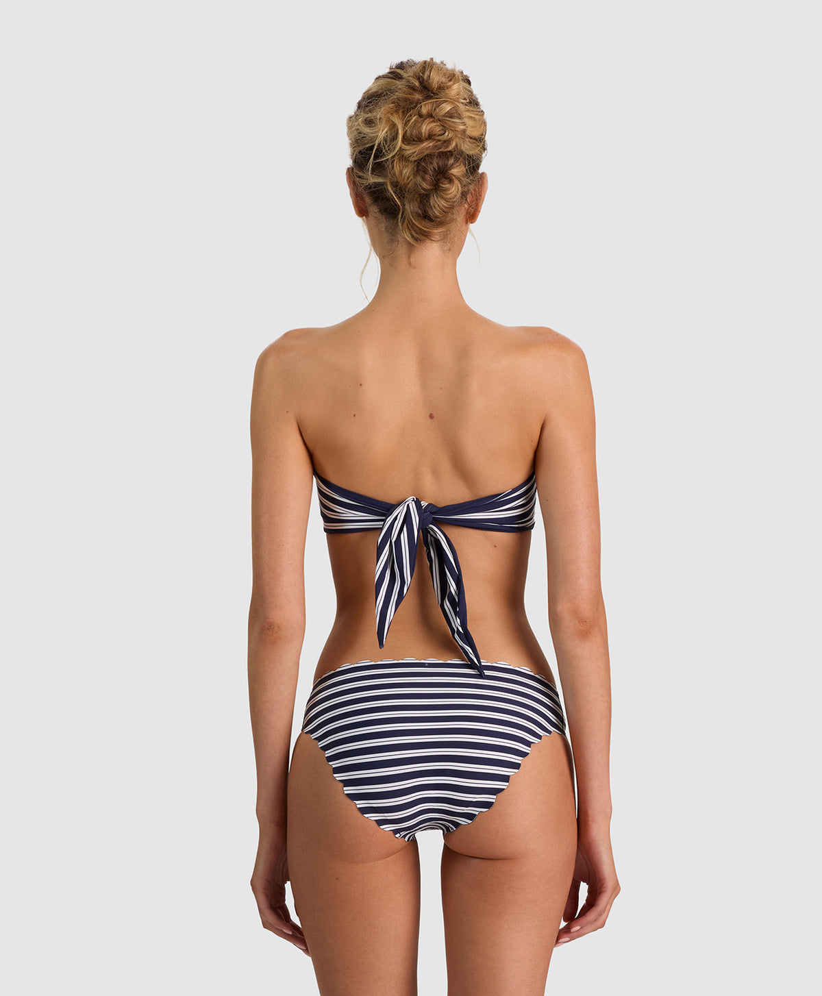A woman viewed from the back wearing a navy and white striped bikini, featuring a tie-back top and high-waisted bottoms, set against a solid light gray background.
