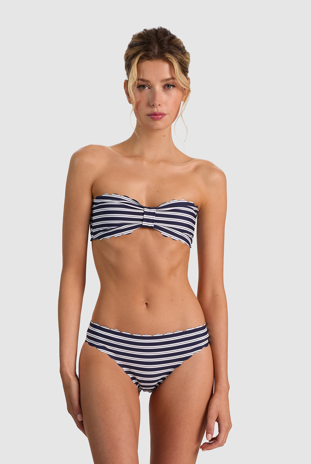 A woman modeling a two-piece bikini with navy blue and white horizontal stripes, featuring a twisted bandeau-style top and high-waisted bottoms, against a solid light gray background.