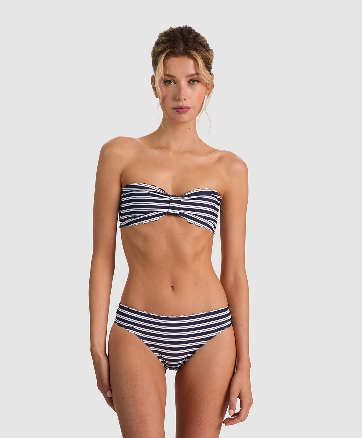 A woman modeling a two-piece bikini with navy blue and white horizontal stripes, featuring a twisted bandeau-style top and high-waisted bottoms, against a solid light gray background.