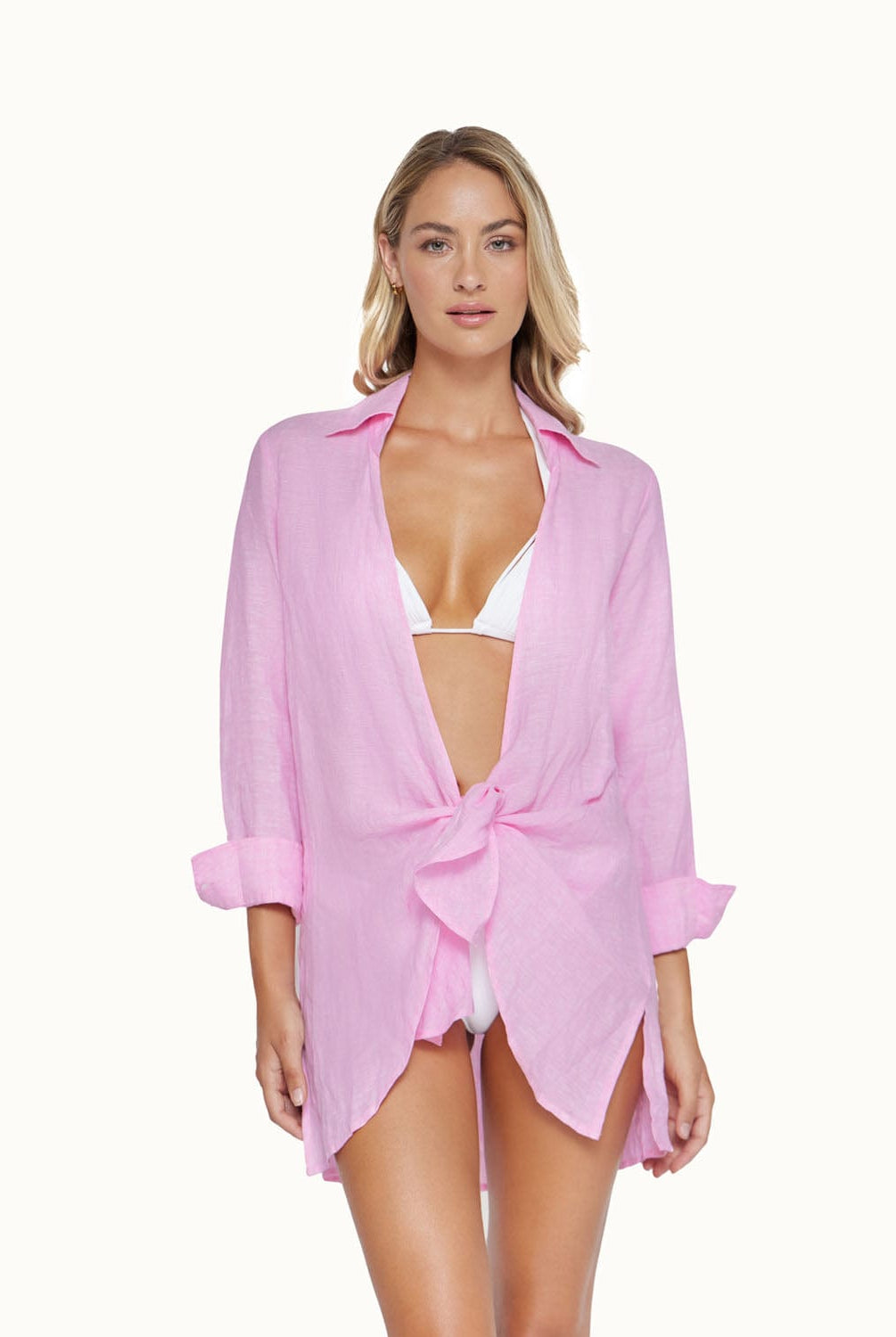 A blonde woman wearing a white bikini and long sleeve pink cover up standing in front of a white wall.