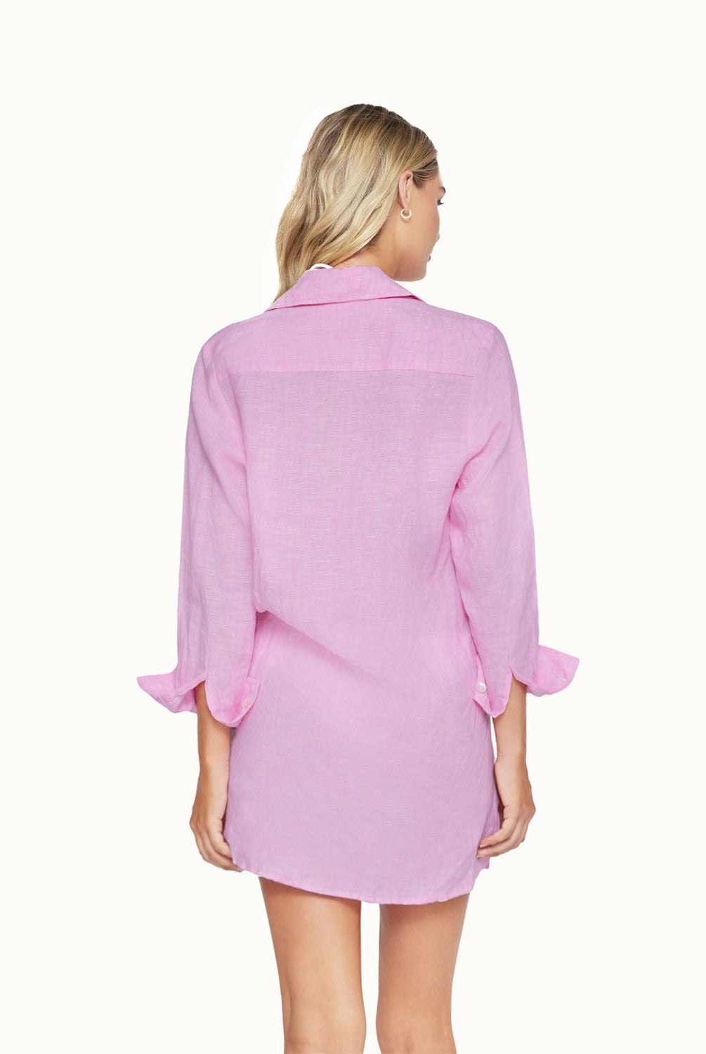 A blonde woman wearing a long sleeve pink cover up facing towards a white wall.