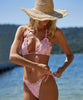 A blonde woman wearing a pink lace bikini and shorts standing near the water.
