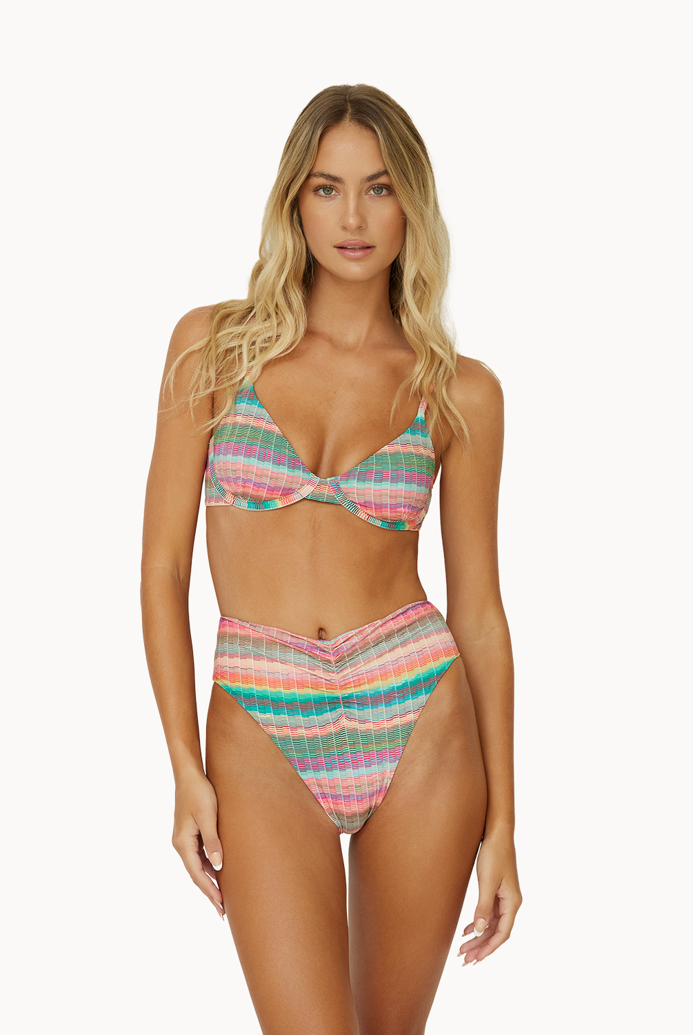 A blonde woman wearing a striped bikini standing against a white wall.