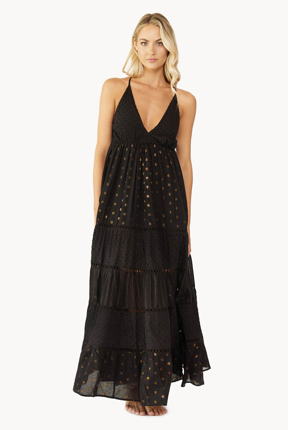 Blonde woman wearing a black full length dress with shimmery gold accents and cross stitching embroidery stands in front of white wall.