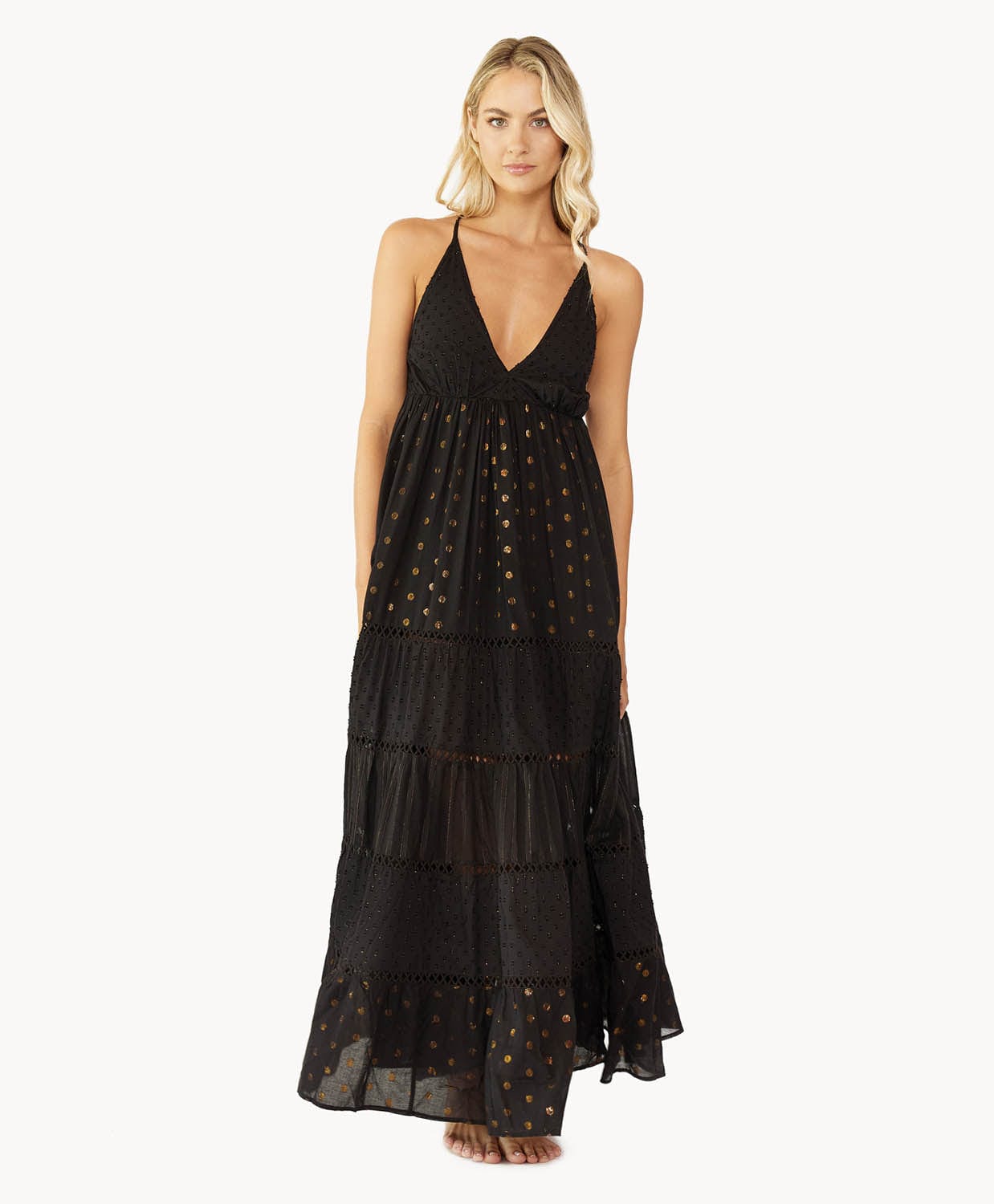Blonde woman wearing a black full length dress with shimmery gold accents and cross stitching embroidery stands in front of white wall.