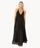Blonde woman wearing a black full length dress with shimmery gold accents and cross stitching embroidery stands in front of white wall.