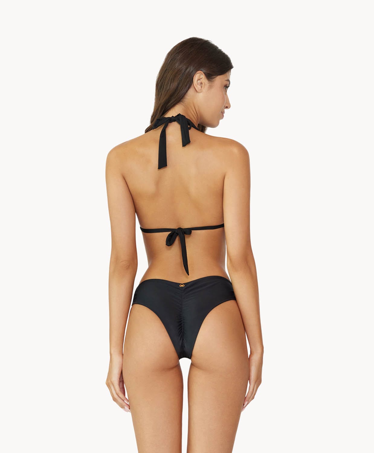 Brunette woman wearing a black ruched bikini facing backwards towards a white wall.