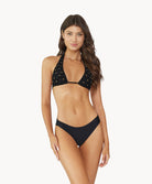Brunette woman wearing a black halter bikini with net macramé details & shimmery gold accents standing in front of white wall.