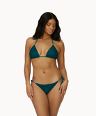 A woman wearing a green triangle top bikini standing in front of a white wall.