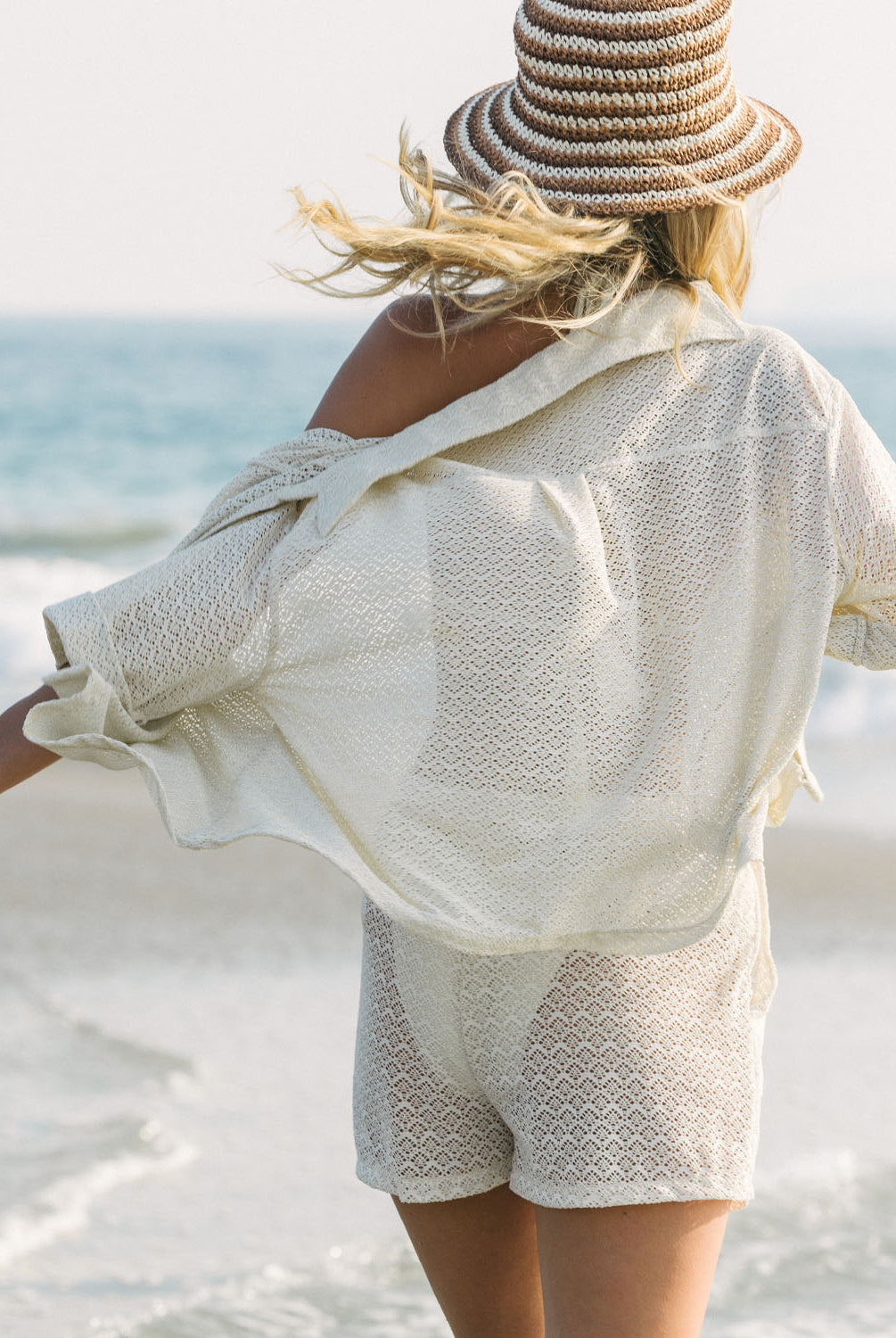 Woman in a beige outfit and straw hat standing on a beach with ocean view