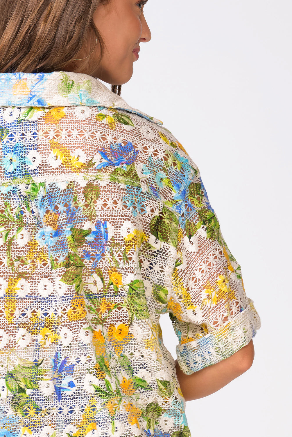 A woman wearing a floral crochet shirt facing towards a white wall.