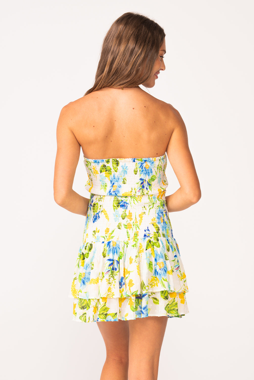 A woman wearing a short strapless dress with a floral pattern facing towards a white wall.
