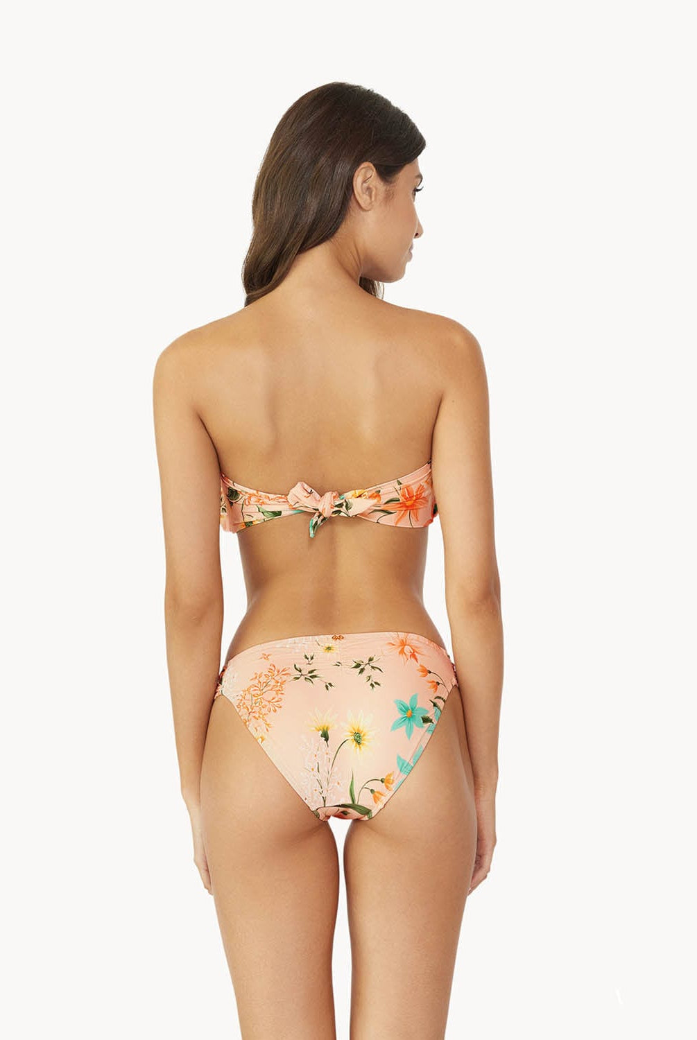 A brunette woman wearing a bikini with a tropical print facing a white wall.