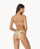 A brunette woman wearing a bikini with a tropical print facing a white wall.