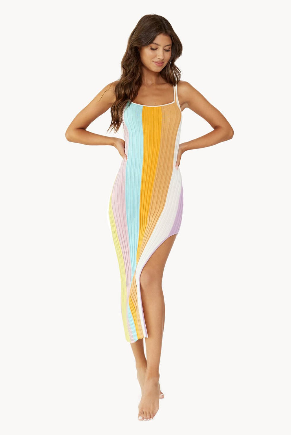 A brunette woman wearing a multi stripe dress stands in front of a white wall.