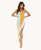 A brunette woman wearing a multi stripe dress stands in front of a white wall.