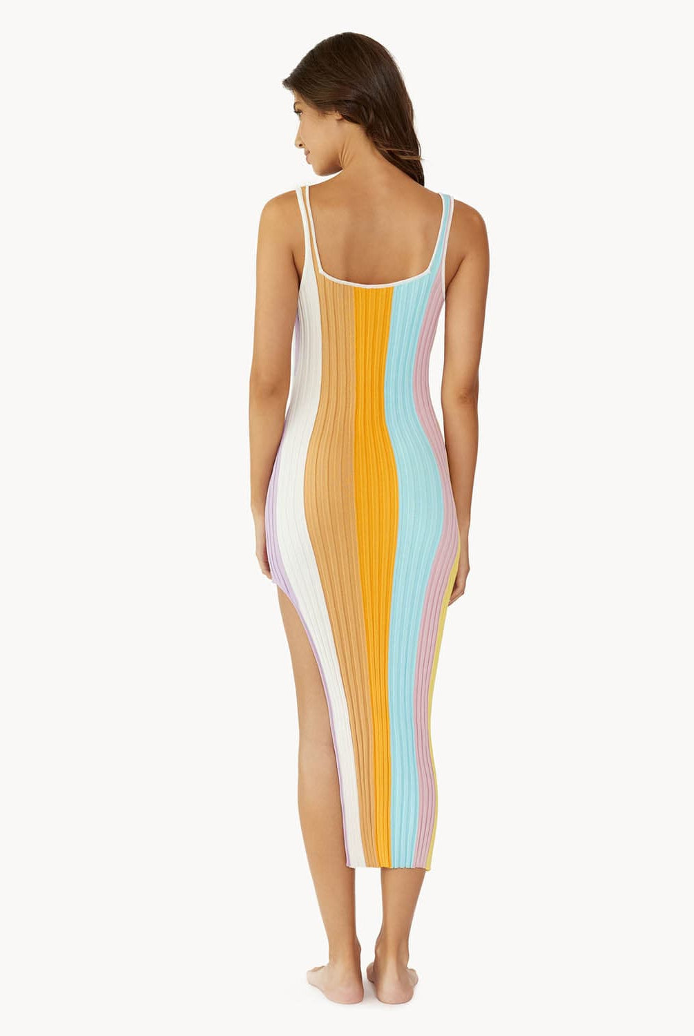 A brunette woman wearing a multi colored stripe dress facing towards a white wall.