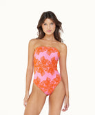 A brunette woman wearing a pink & orange one piece suit standing against a white wall.