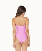 A brunette woman wearing a pink one piece suit facing towards a white wall.