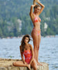 A brunette woman wearing a pink & orange one piece next to a blonde woman wearing a pink & orange bikini. Both woman standing on a rock near the water.
