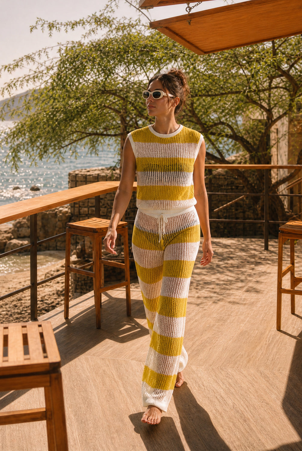 A woman wearing a matching yellow and white striped knit two-piece set walks barefoot on a wooden deck with ocean and trees in the background.