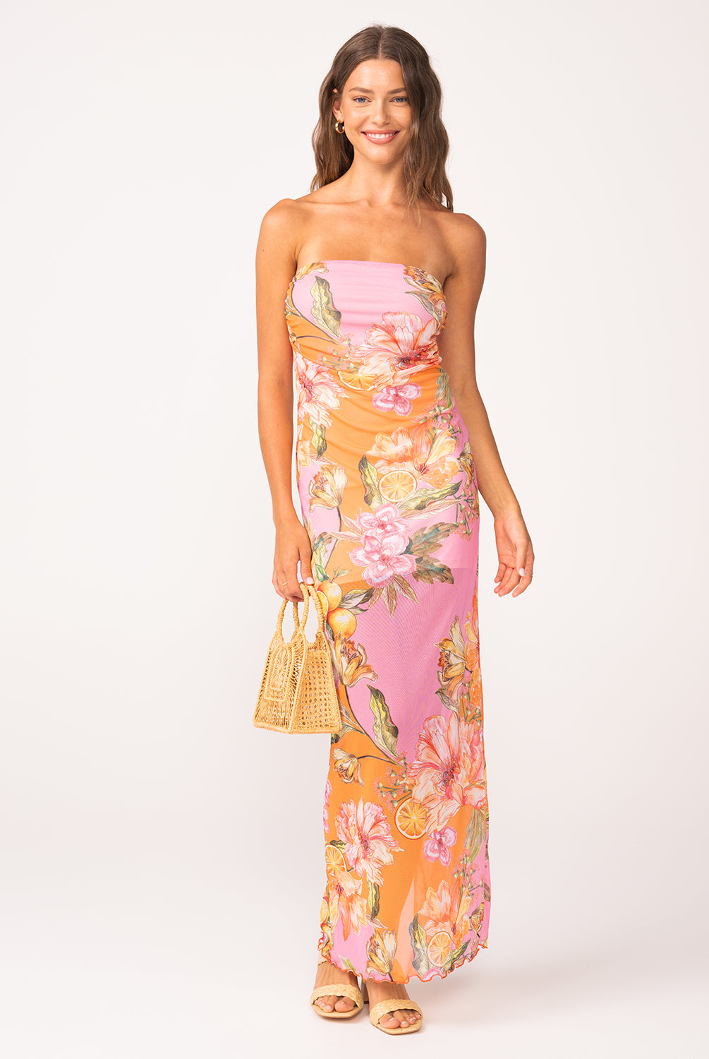 A woman wearing a long, strapless, pink and orange dress with a floral print standing against a white wall.