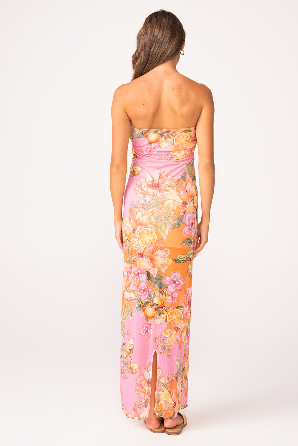 A woman wearing a long, strapless, pink and orange dress with a floral print facing towards a white wall.