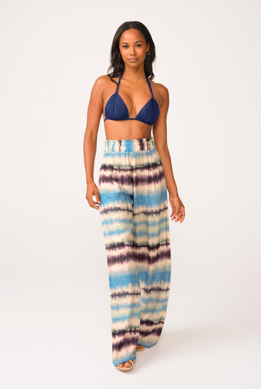 A woman wearing a navy triangle bikini top and blue tie dye pants standing against a white wall.