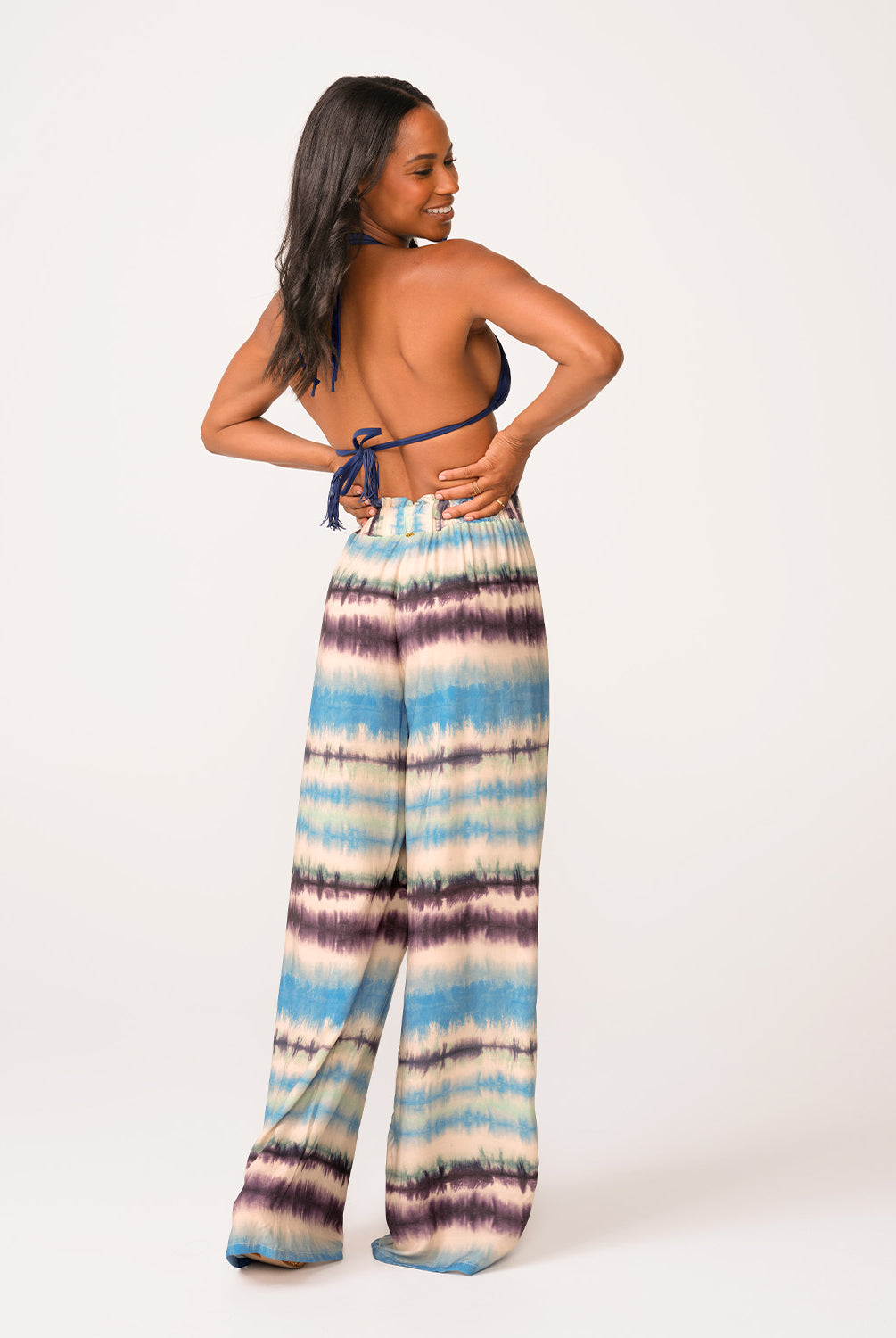 A woman wearing a navy triangle bikini top and blue tie dye pants standing against a white wall.