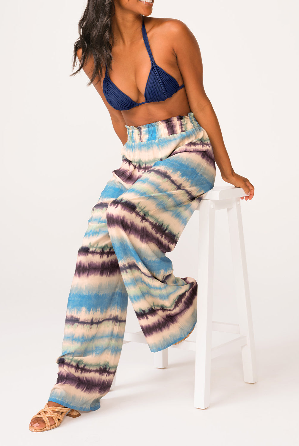 A woman wearing a navy triangle bikini top and blue tie dye pants sitting on a white stool.