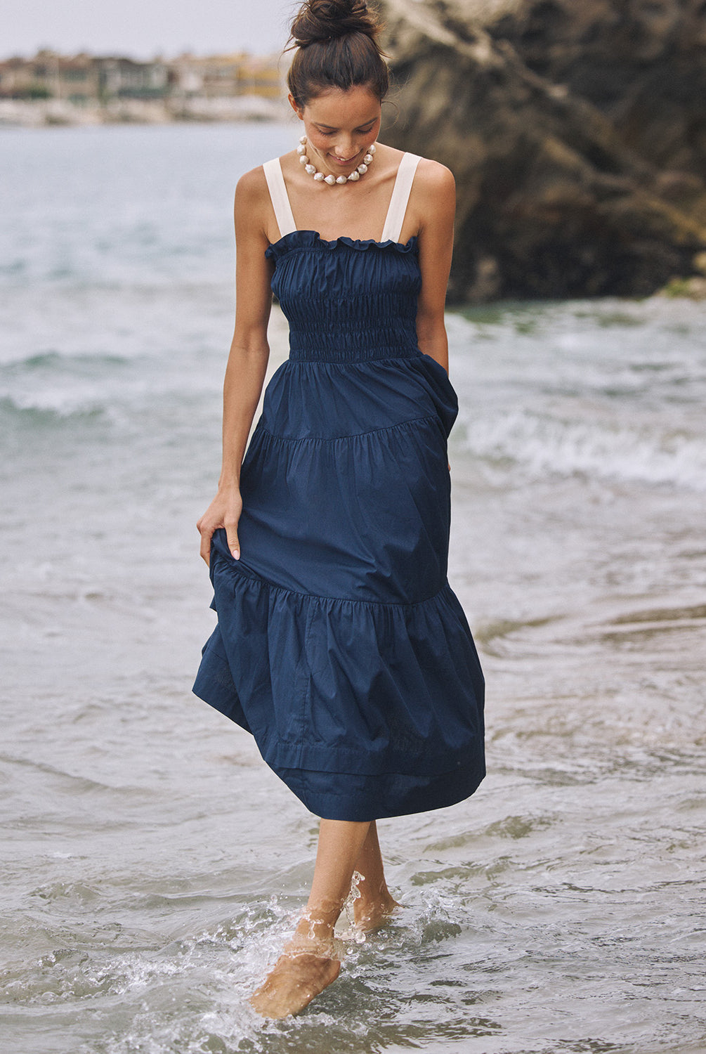 A woman wearing a navy dress with white straps walking in the ocean.