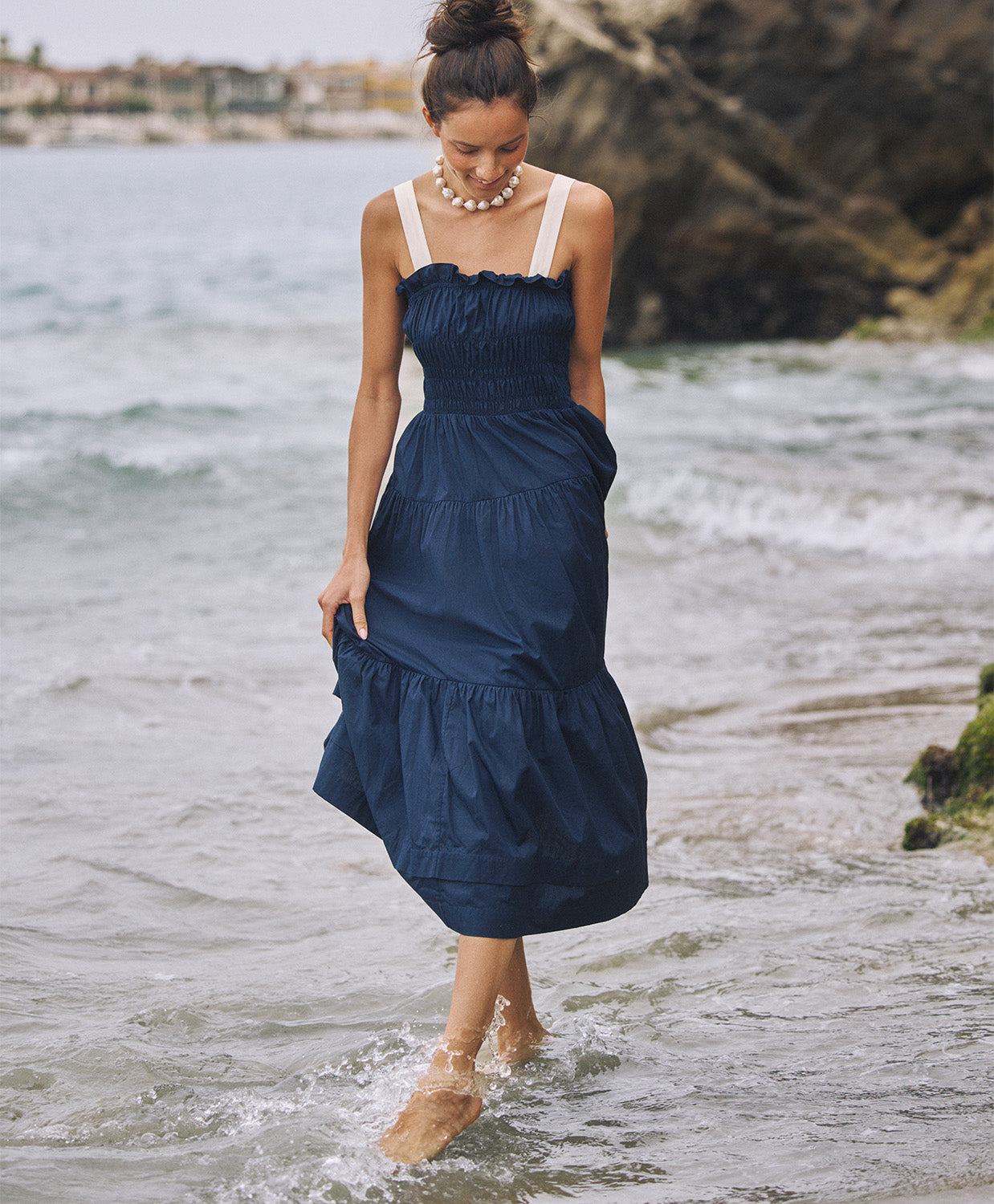 A woman wearing a navy dress with white straps walking in the ocean.