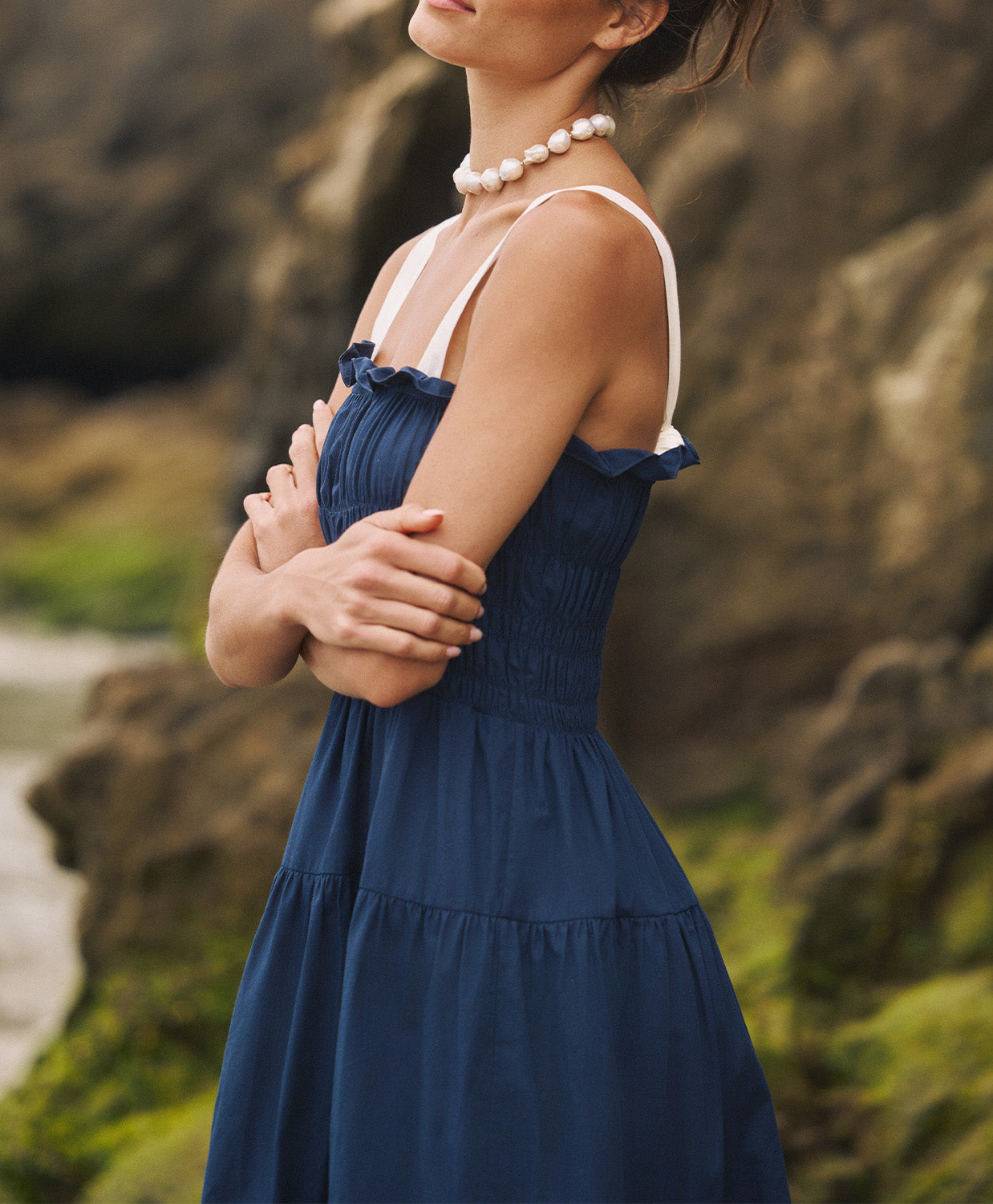 A woman wearing a navy dress with white straps standing outside near a rock wall.