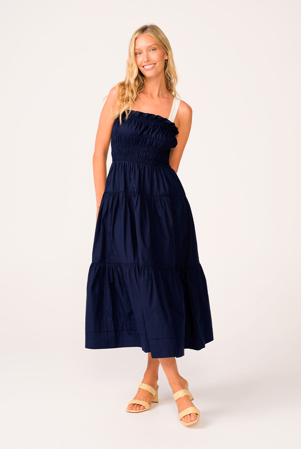 A woman wearing a navy dress with white straps standing against a white wall.