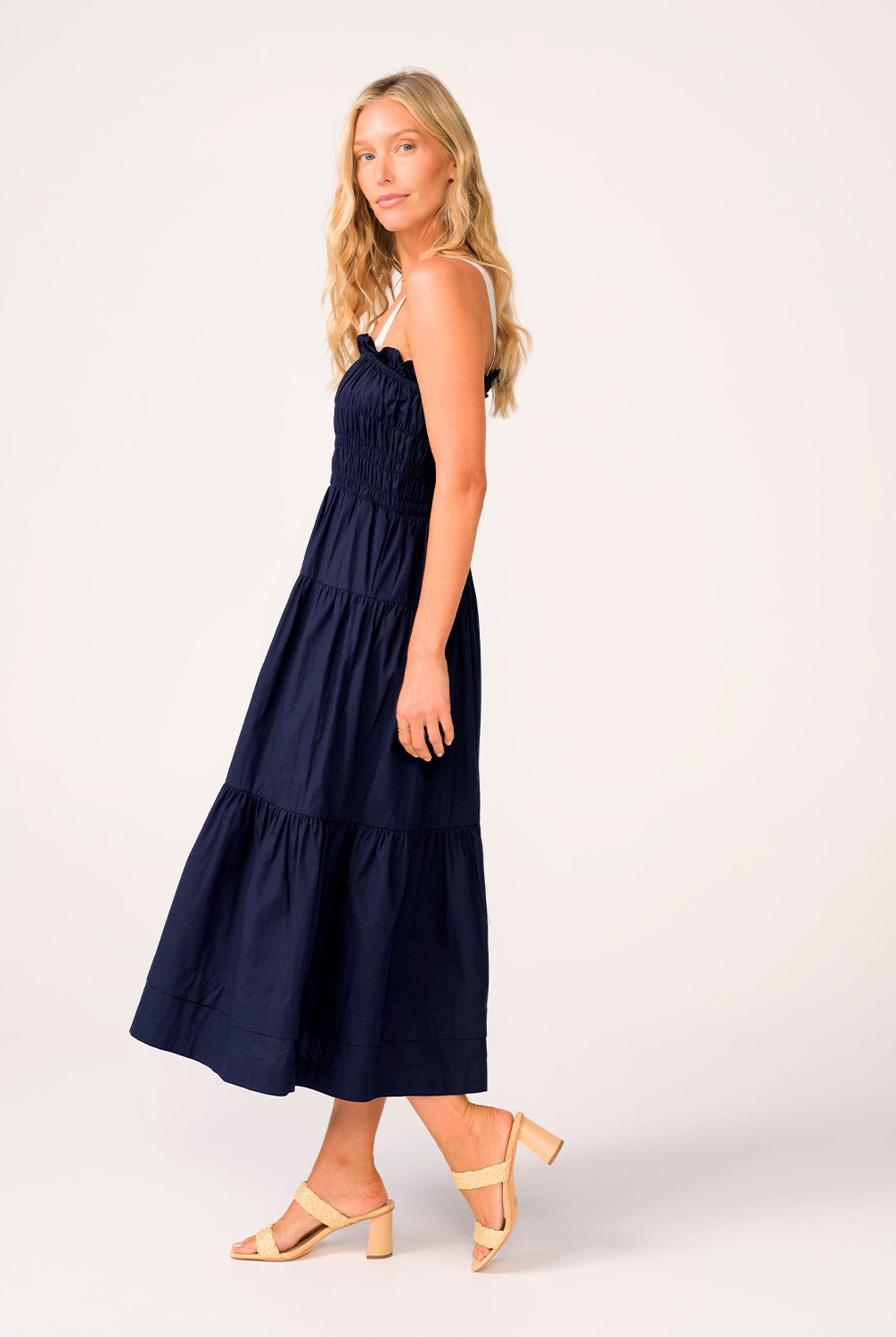 A woman wearing a navy dress with white straps standing against a white wall.