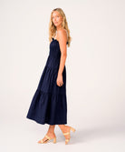 A woman wearing a navy dress with white straps standing against a white wall.