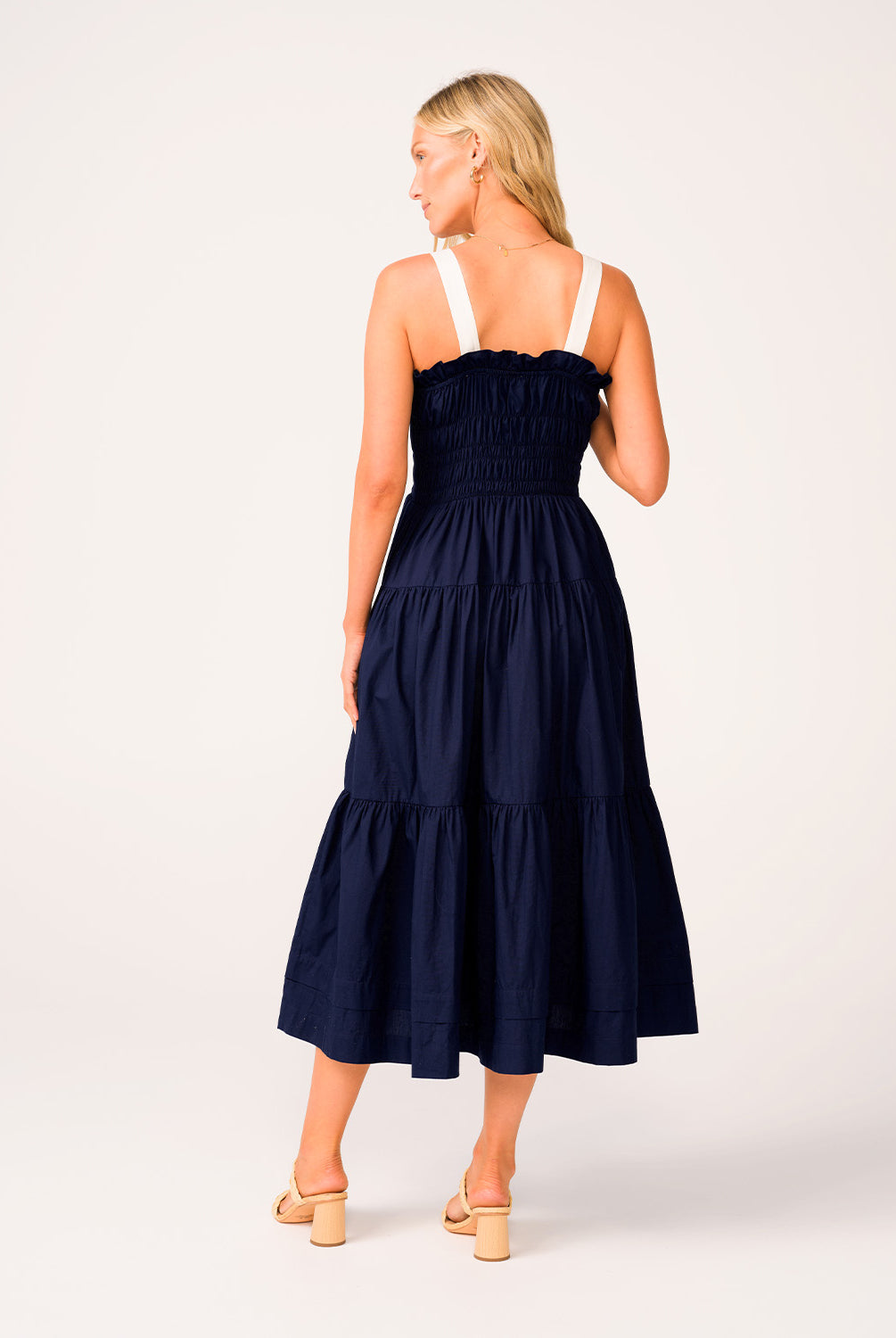 A woman wearing a navy dress with white straps standing against a white wall.