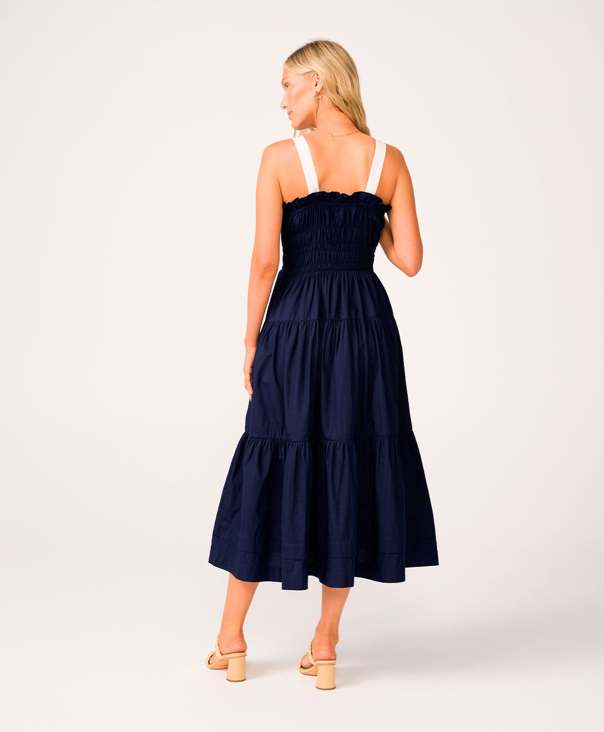 A woman wearing a navy dress with white straps standing against a white wall.