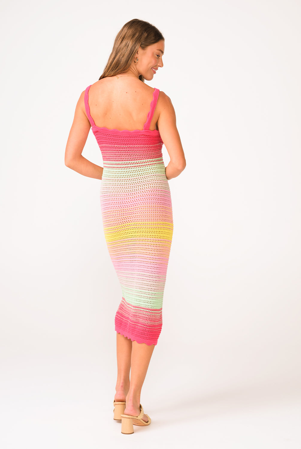 A woman wearing a pink, green, and yellow crochet knit mid length dress facing towards a white background.